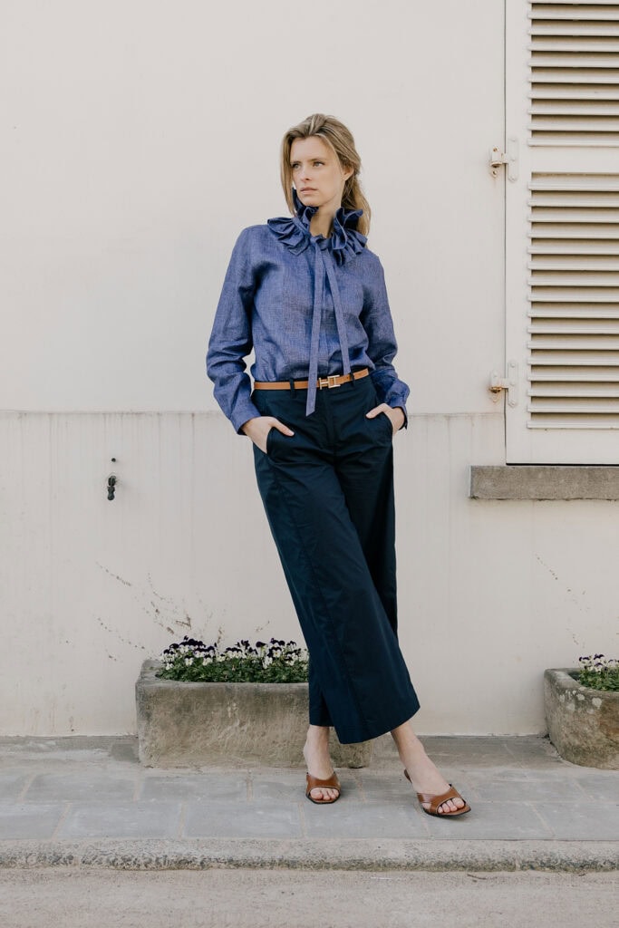 Vrouw in blauwe blouse en donkere broek, staand buiten tegen witte muur.