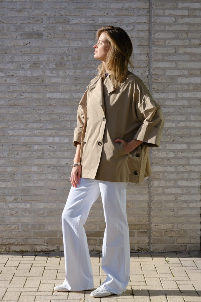 Vrouw in beige jas en witte broek poseert tegen bakstenen muur.