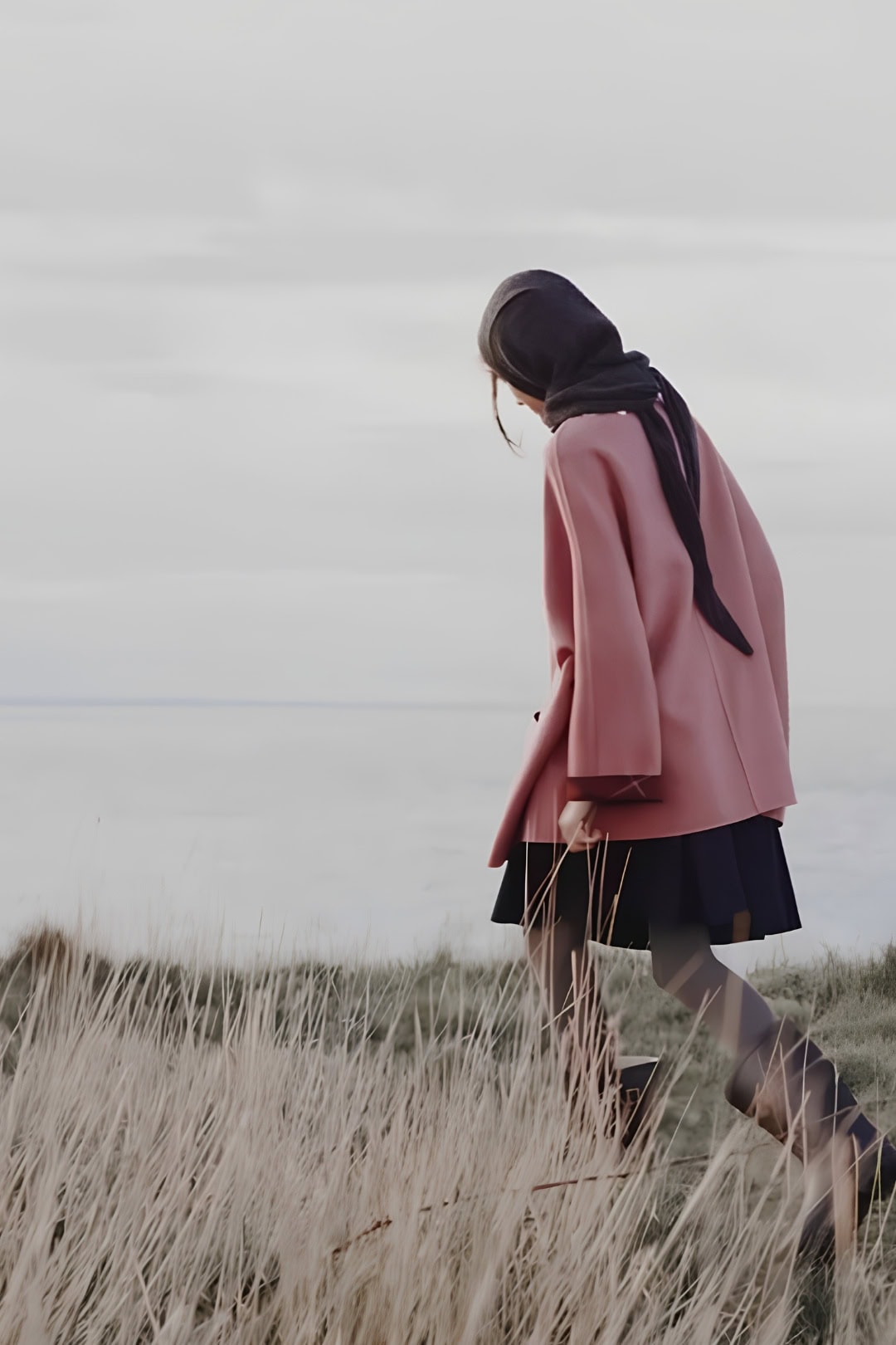 Vrouw in roze jas en hoofddoek wandelt door grasland bij zee.