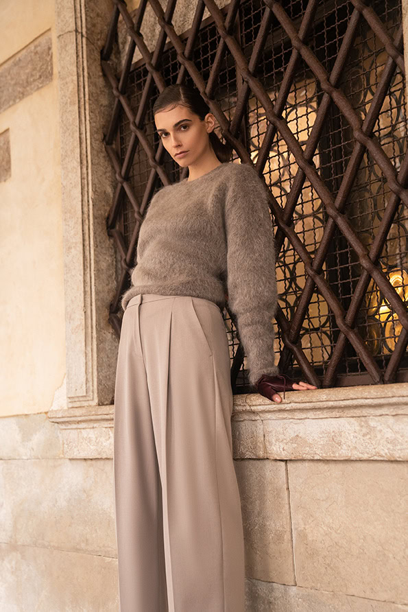 Modieuze vrouw in grijze trui en beige wijde broek bij historische gevel.