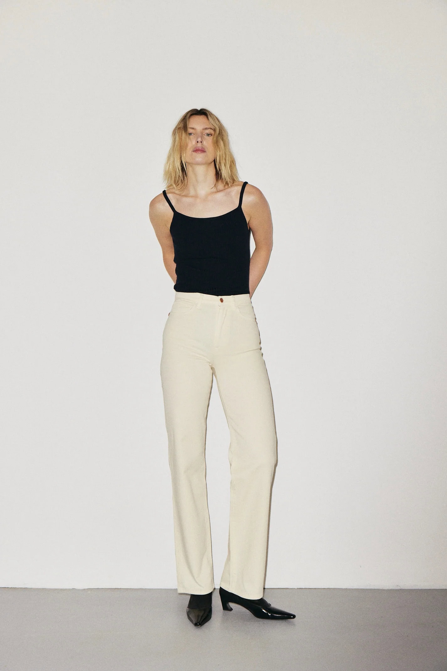 Modieuze vrouw in zwart top en witte broek op witte achtergrond.